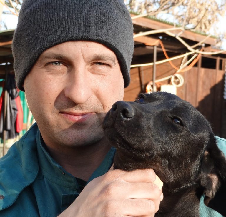 Mann mit schwarzem Hund, beide schauen in die Kamera, im Hintergrund eine Gartenlandschaft.